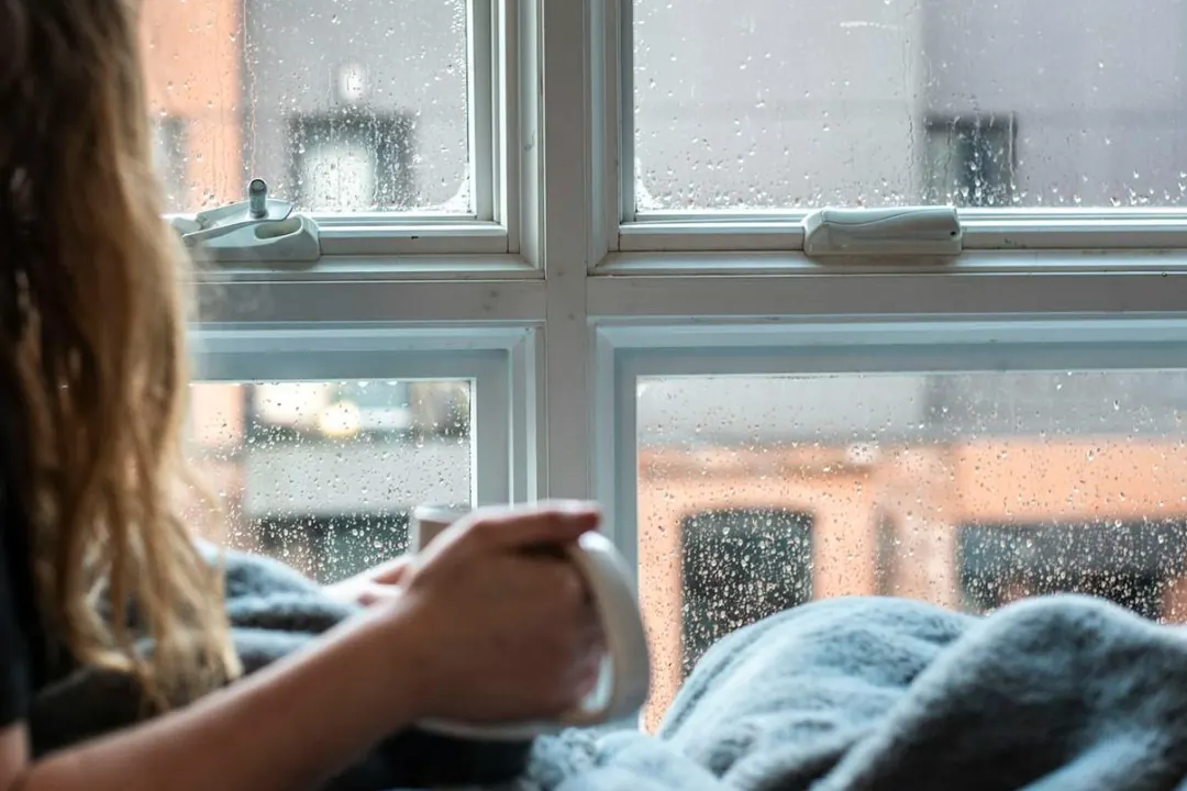 Hands holding a warm mug beside a rainy window, a soft blanket in the foreground